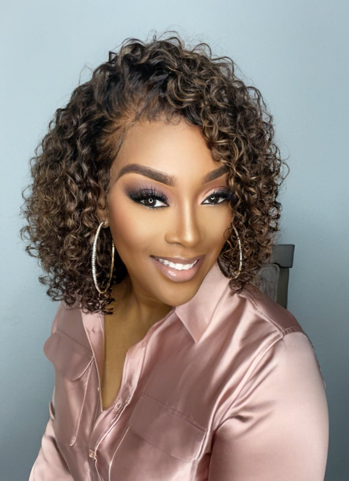 A woman wearing a raw curly highlighted dyva cut HD lace bob wig in a cinnamon brown color with deep curly texture.